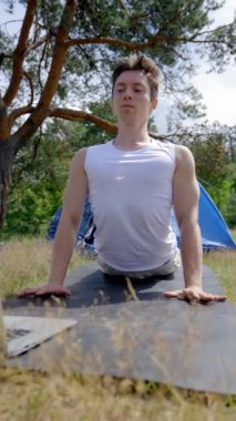 Siyah saçlı kampçı laptopta siyah hasırın üzerinde egzersiz yapıyor. Genç adam internetten yoga videosu buluyor ve kamp çadırının yakınında gökyüzüne karşı dikey çekim yapmaya karar veriyor..