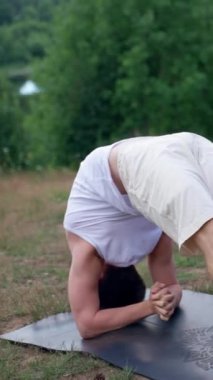Gençler çimenli tepedeki minderlerde birlikte yoga yapıyorlar. Konsantre bir erkek ve kadın tekerlek pozu verir ve doğayla birleşir..