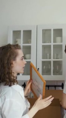 Genç çift, yeni daireye taşındıktan sonra karton kutulardan bir şeyler çıkarıyor. Mutlu kadın, fotoğrafı ahşap çerçeveye koyar mutfaktaki rafta dikey çekim.