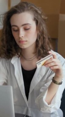 Genç bir kadın, kart detaylarını girerek online mağazadan mutfak gereçleri alıyor. Ev hanımı paketlenmiş kağıt kutuların karşısında oturuyor. Dikey çekim.