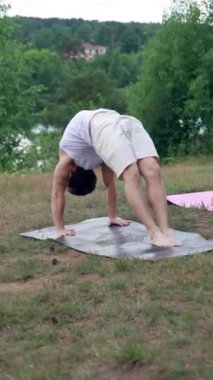 Birkaç genç, nehir kıyısındaki minderlerde yoga tekeri pozu veriyor. Rahat elbiseli kadın ve erkek bulutlu havada doğayla birleşir..