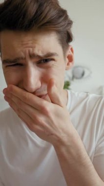 Şok olmuş, suratını buruşturan ve kameraya bakmayı reddeden genç bir adam. Modern oturma odasında oturan adam video görüşmesinde arkadaşlarıyla dikey çekim yapıyor..