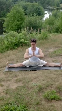 Genç kaslı adam, namaste ellerle katlanmış ve açık havada bacaklarını açan gözleri kapalı bir şekilde mindere oturur. Atletik adam yüksek nehir kıyısında yeşil ağaçlara karşı pratik yapıyor..