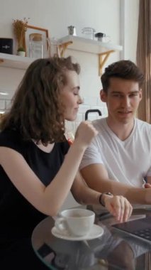 Genç nişanlı çift, dizüstü bilgisayarla arkadaşlarıyla düğün hakkında konuşuyor. Mutlu adam ve kadın mutfak masasında oturur ve dikey kamera görüntülerine bakarlar..