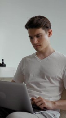Genç adam beyaz duvara karşı sandalyede oturuyor ve şık bir odadan dizüstü bilgisayarla çalışıyor. Yeni projenin erkek serbest yazar tipleri modern elektronik aygıt Dikey Vuruş kullanılarak rapor edilir.