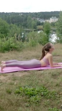 Genç kadın çimenli tepede yoga pozu veriyor. Pembe spor giysili atletik kadın, Asana Upward-Facing Dog 'a nehrin kıyısındaki yeşil ormana karşı konsantre oluyor..