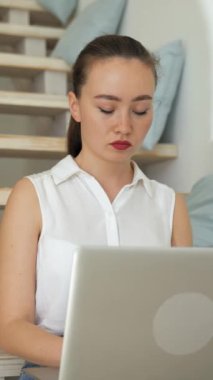 Genç bir kadın dizüstü bilgisayarda çalışır ve kameraya döner ve gülümser. Serbest çalışan kadın, modern cihazın dikey vuruşu üzerine makale yazarak tahta merdivenlerde oturuyor..