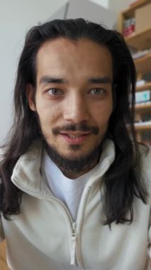 İnternetteki iş konferansında kamerayla konuşan uzun saçlı genç adamın görüntüsü. Çalışan, raflara karşı işyerinde dikey çekim yapan ürünlerle oturuyor.