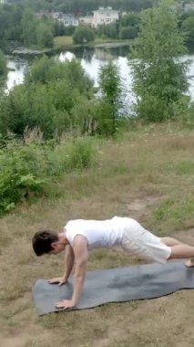 Genç atletik adam çimenli çayırlarda yoga pozisyonu sergiliyor. Konsantre olmuş, rahat giysiler içindeki adam karnının üzerine uzanmış ellerini paspasın üstüne koyup bacaklarını tutarak dikey çekim yapıyor..