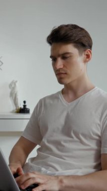 Odaklanmış çalışan dizüstü bilgisayardaki yazı ekranına bakar. Genç adam internetten modern elektronik cihazla çalışıyor. Oturma odasındaki sandalyeye oturtuluyor. Dikey çekim..