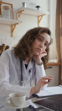 Uykulu kadın pratisyen dizüstü bilgisayarla sıkıcı eğitici konferansları dinliyor. Üniformalı genç bir kadın mutfakta cam masada oturmaktan yoruldu..