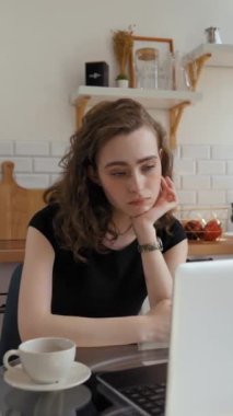 Yorgun bir kadın, mutfaktaki cam masada oturup dizüstü bilgisayarla eğitim veren kurslar dinliyor. Genç kız öğrenci evde çalışırken uykulu hissediyor..