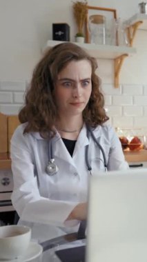 Genç bayan doktor, dizüstü bilgisayardaki çevrimiçi video kurslarında tatsız anlara bakıyor. Kadın, evde, mutfakta, dikey çekimde öğrenim kalitesini artırıyor..