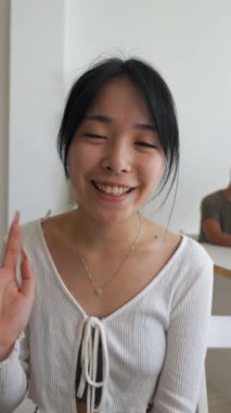 Neşeli Asyalı kadın görüntülü telefonda arkadaşıyla konuşuyor ve uzaklaşmak istiyor. Genç bayan işyerinde oturuyor. Yakın plan dikey çekim..