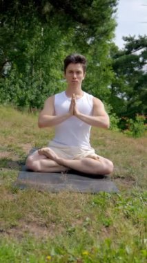 Genç atletik adam Lotus 'ta meditasyon yapıyor. Namaste' nin çimenli çayırlarda gözlerini kapamasıyla. Rahat elbiseli adam mindere oturup temiz hava soluyor. Dikey çekim..