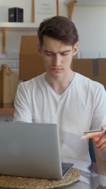 Genç adam, mutfaktaki karton kutuların yanında oturan dizüstü bilgisayarla online alışveriş yapıyor. Adam yeni ev için ev aletleri alıyor kart detaylarını dikey olarak giriyor.