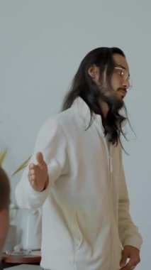 Gözlüklü erkek antrenör, seyirciler arasında masada oturan öğrencilerle iletişim kurmaktan hoşlanıyor. Uzun saçlı genç adam yavaş çekim dikey çekim yapan insanlarla konuşurken güler..