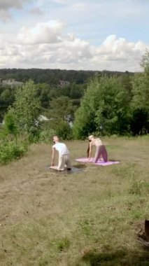 Genç bir adam ve kadın dik nehir kıyısındaki minderlerde yoga devesi pozu veriyorlar. İnsanlar Asana 'ya konsantre olup yüksek tepelerde dikey çekim yapıyorlar..