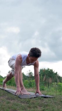Genç adam yeşil tepede ormana karşı yoga yapıyor. Atletik Yogi, ufukta bulutlu gökyüzüne karşı Asya Aşağı Yüzlü Köpek üzerinde yoğunlaşır..