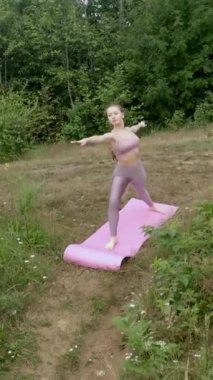 Genç bir kadın, dik nehir kıyısında yoga savaşçısı pozu veriyor. Yoğunlaşmış bayan, yeşil ağaçlara karşı yüksek tepelerde eğlenerek, Asana 'yı sahneliyor..