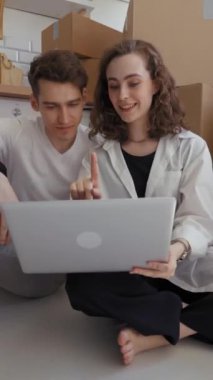 Mutlu kadın, erkek odasının tasarımını mutfakta, yeni dairede karton kutuların yanında gösteriyor. Genç çift dizüstü bilgisayardaki yenilenme fotoğraflarına bakıyor..