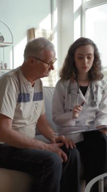 Beyaz önlüklü bayan doktor evde yaşlı bir hastaya reçete yazıyor. Uzman, oturma odasındaki kanepede oturan yaşlı adama tedavi planı gösteriyor..