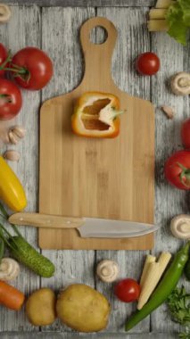 Bir insan eli kesme tahtasından portakallı biberin yarısını alıyor. Ahşap masada vejetaryen yemeği pişirmek için bıçak ve diğer sebzeler. Dikey çekim.