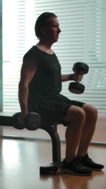 Olgun bir adam pazılarını şişirmek için büyük dambılları kaldırıyor, spor salonundaki spor salonunda tütsülenmiş spor masasında oturuyor. Ağır çekim geniş çekim. Dikey çekim.