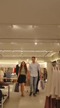 Alışveriş paketleri olan mutlu evli çift kameraya gülümseyerek yaklaşıyor. Yeni, modern bir giyim mağazasından alınmış tişörtlü sevimli bir aile. Orta atış. Dikey çekim.