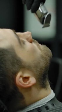 Berber kız müşterilere tıraş için sakal hazırlıyor. Siyah eldivenli, tanınmayan sarışın bir kadının genç bir adamı tıraş edişinin yakın çekimi. Dikey çekim.