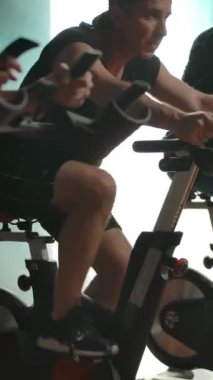 Sporcular spor salonunda peş peşe egzersiz bisikletlerinde bacak kaslarını çalıştırıyorlar. Sağlıklı yaşam tarzı konsepti, gençliği koruma arzusu. Son olarak güzel bayana odaklanıyorum. Dikey çekim.