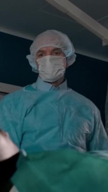 Düşük açılı, üniformalı, koruyucu maskeli ve eldivenli yetişkin erkek cerrahın hastane ameliyathanesinde tıbbi prosedüre hazırlanırken el kaldırması. Dikey çekim.
