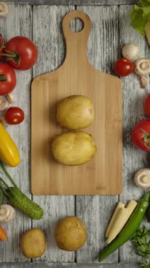 Kesme tahtasının üzerinde iki patates meyvesi var. Erkek eli patatesi aynı anda alıyor. Vejetaryen yemeği pişirmek için sebzeli kırsal bir çelik aşçının dikey görüntüsü. Dikey