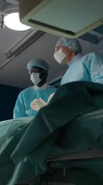 İki cerrah ameliyat için bir röntgen cihazını hareket ettiriyor. Maskeli, cüppeli, esmer bir doktor ve olgun meslektaşı hastayı ameliyata hazırlıyor. Dikey çekim.