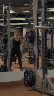 Rear view of sexy caucasian woman trainer doing barbell squats indoor of gym. Good exercises to build a muscular butt. Modern sport hall interior. Vertical shot.