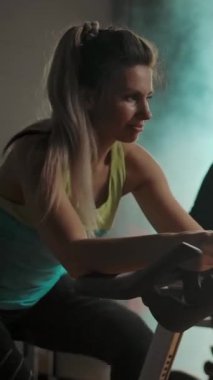 Sportif sporcular, iki erkek ve bir kadın bisiklet simülatörü, dönen pedallar. Modern spor salonunda sağlıklı bir yaşam tarzı. Yavaş çekim takip çekimi. Taytlı seksi kadına odaklan. Belirsiz arkaplan