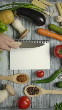 Ahşap tezgahın üzerine malzemeler ve çatal bıçaklarla yerleştirilmiş beyaz boş bir sayfa ile yemek kitabına işaret eden aşçı manzarası. Makbuzunu boş bir kağıda koymak kolay. Dikey çekim.