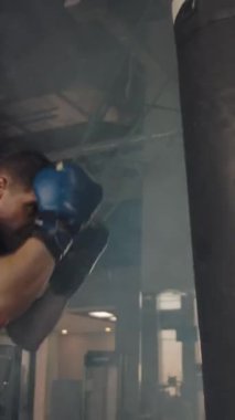 Genç profesyonel atlet eldiven giyerken sert bir şekilde kum torbasına vuruyor. Egzersiz aletleri olan dumanlı spor salonu. Dikey çekim.