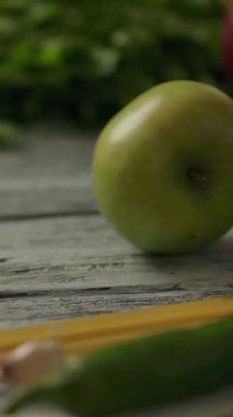 Tanınmayan biri, organik meyve ve sebzelerin arasında ahşap kırsal masada taze yeşil elma itiyor. Dikey çekim.