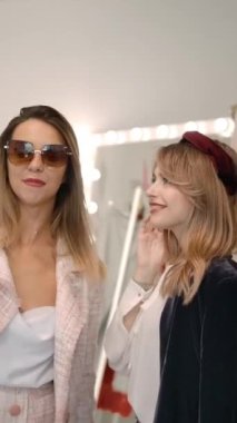Şık takım elbiseli, şık güneş gözlüklü modern şık sarışın kadın akıllı telefon kullanıyor ve moda blogu için video kaydediyor. Bir yandan da mağaza görevlisi ile butikte dikiliyorlar. Dikey çekim.