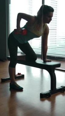 Diz çökmüş spor bankları üzerinde triseps egzersizi yapan genç bir kadın. Kamera seksi kadının etrafında dönüyor. Modern jimnastik salonu, yavaş çekim orta çekim. Dikey çekim.