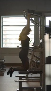 Koç kıza gravitron üzerinde nasıl egzersiz yapacağını tavsiye etti. Genç, güzel kızıl saçlı kadın spor salonundaki modern egzersiz makinesinde barfiks çekiyor. Dikey çekim.