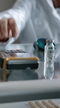 Ödeme kablosuz terminali ve aşı ampulü, kredi kartıyla alınıyor. İçi ilaç, hap ve ampul dolu raflarla dolu bir eczane. Dikey çekim.