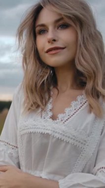 Kamera, bulutlu gökyüzüne karşı yeşil çayırda dinlenirken, nazik elbiseli, gülümseyen ve kameraya bakan romantik yalnız bir kadının etrafında dönüyor. Dikey çekim.