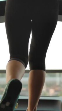 Spor salonunda koşu bandında güzel seksi kadın bacakları. İnce, beyaz, sarışın bir kızın dikey çizimi koşu bandında koşup pencereden dışarı bakıyor. Yavaş çekim dikey çekim.