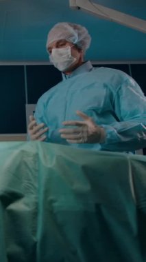 Üniformalı ve maskeli bir erkek cerrahın kadın hastayla konuşması ve modern ameliyathanede ameliyat öncesi prosedürü açıklaması. Dikey çekim.