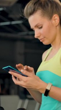 Modern bir spor salonunda koşu bandında yürürken bir mesaj yazan ve cep telefonuyla sohbet eden güzel beyaz bir kızın portresi. Dikey çekim.