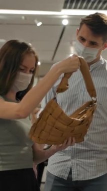 Sağlık maskeli genç bir adam ve kadın salgın sırasında modern mağazayı ziyaret ederken çantalarını inceliyor. Dikey çekim.