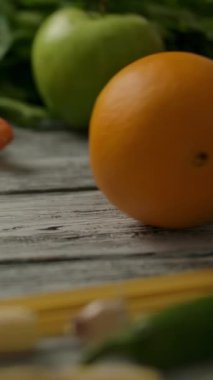 Olgun portakal ve taze elma mutfaktaki çeşitli sebzelerin yanında ahşap masada yuvarlanıyor. Dikey çekim.