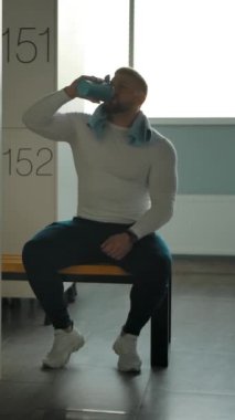 Eğitimden sonra bir adam soyunma odasında su ve havlu içiyor. Güç yüklemesinden dinlenin. Arka planda pencere ve dolaplar var. Yavaş çekim çekim. Dikey çekim.